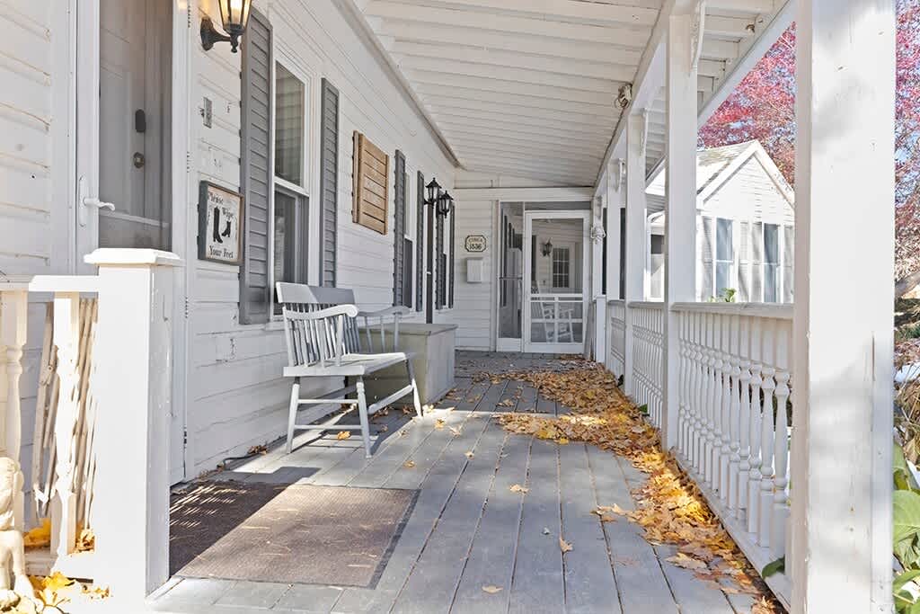 Charming white wooden porch with a railing, a bench, and fallen autumn leaves scattered on the floor. Multiple windows with dark shutters and exterior lights are visible.