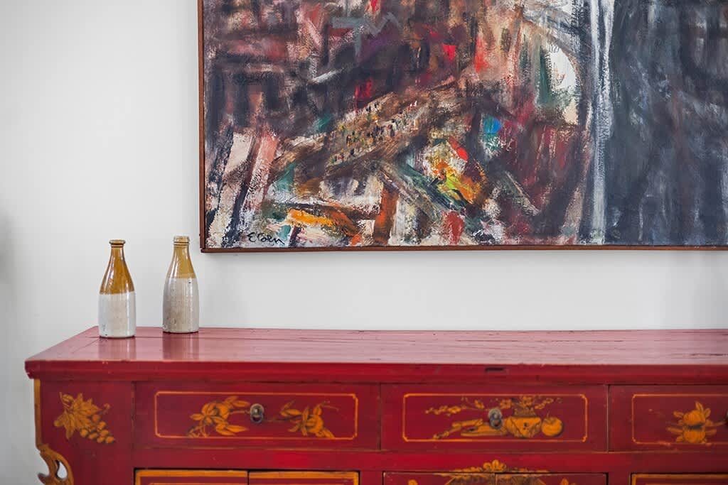 Close-up of a vibrant red console table with intricate gold detailing, positioned against a white wall. An abstract painting with dark, rich colors hangs above the table, flanked by two decorative bottles.