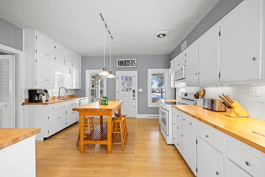 Another view of the bright kitchen, showing white shaker cabinets, light wood countertops, and stainless steel appliances. A wooden island with stools is in the foreground, and a coffee maker and toaster are visible.