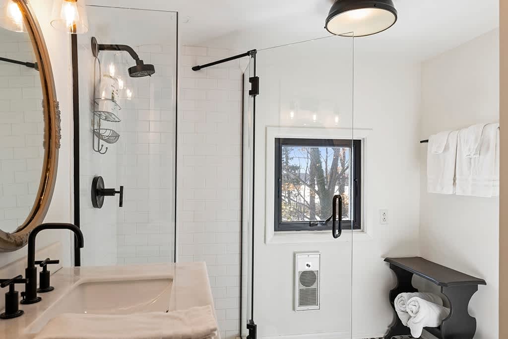 Modern bathroom featuring a glass-enclosed shower with white subway tiles, a dark vanity with a white sink, and a large oval mirror. Black fixtures and a small window add contrast.