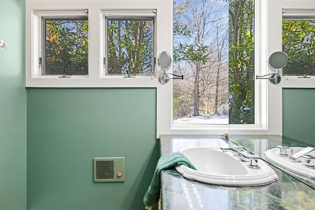 A close-up of the bathroom vanity area, showcasing a white sink set into a green marble countertop, with a large window offering a view of lush trees. A small wall heater is visible below the window.