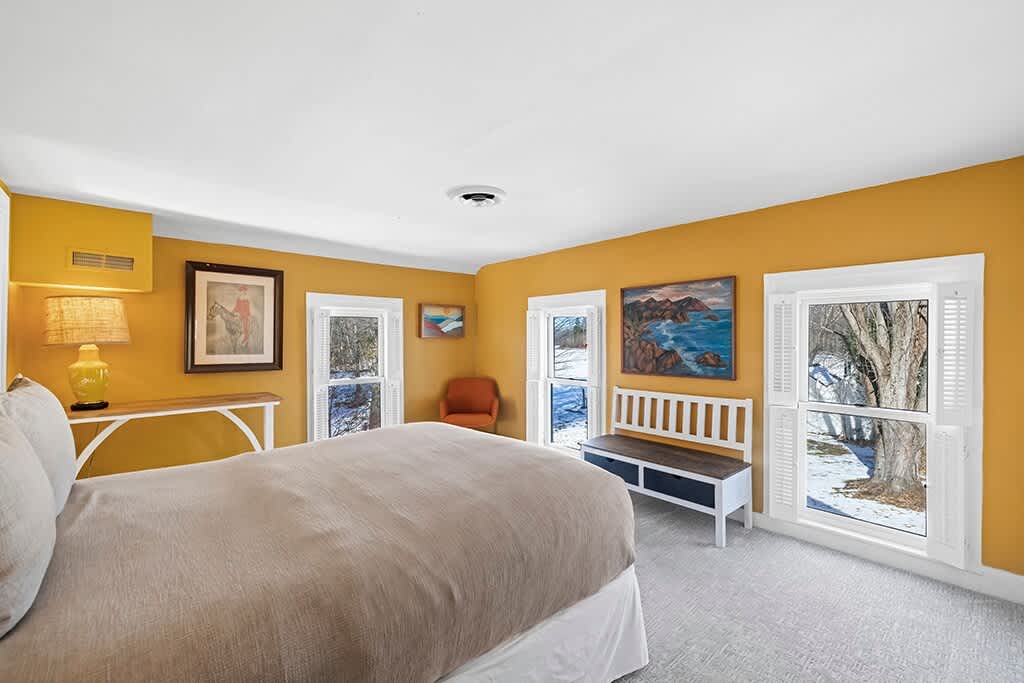A cozy bedroom with mustard yellow walls, a large bed with a beige duvet, and multiple windows offering views of a snowy outdoor scene. Artwork adorns the walls, and a small orange armchair adds a pop of color.