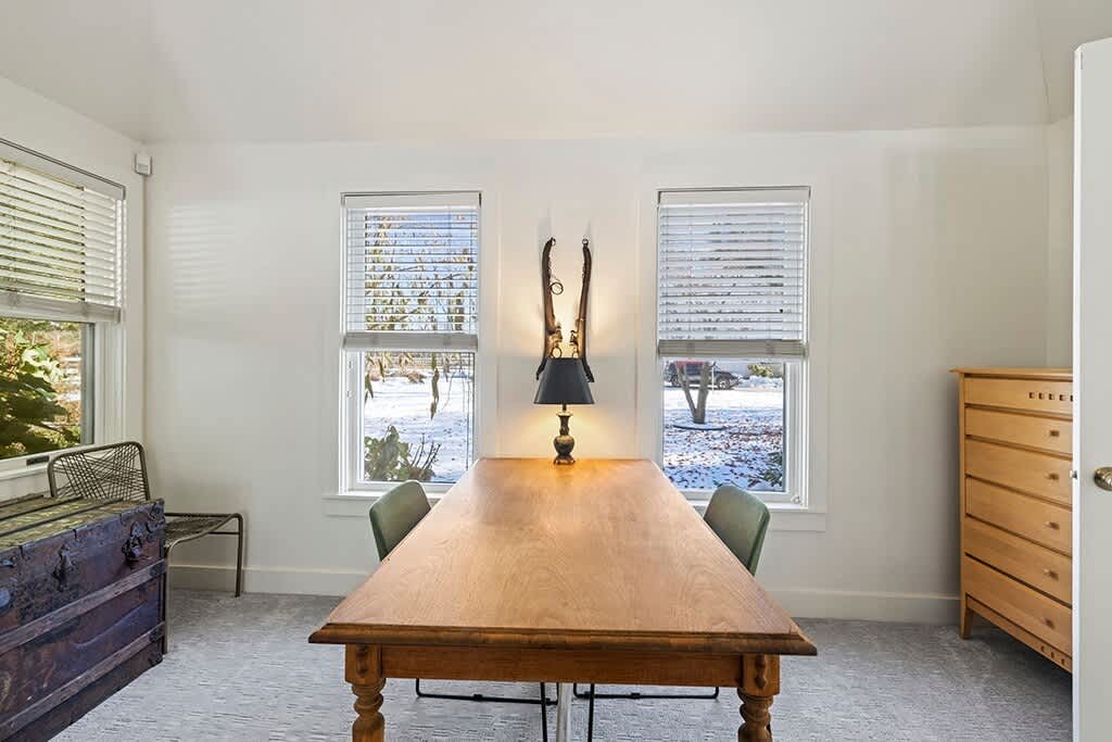 A bright dining room featuring a long, light wood table with two chairs, flanked by two windows overlooking a snowy landscape. A unique wall-mounted decorative piece with a lamp hangs between the windows.