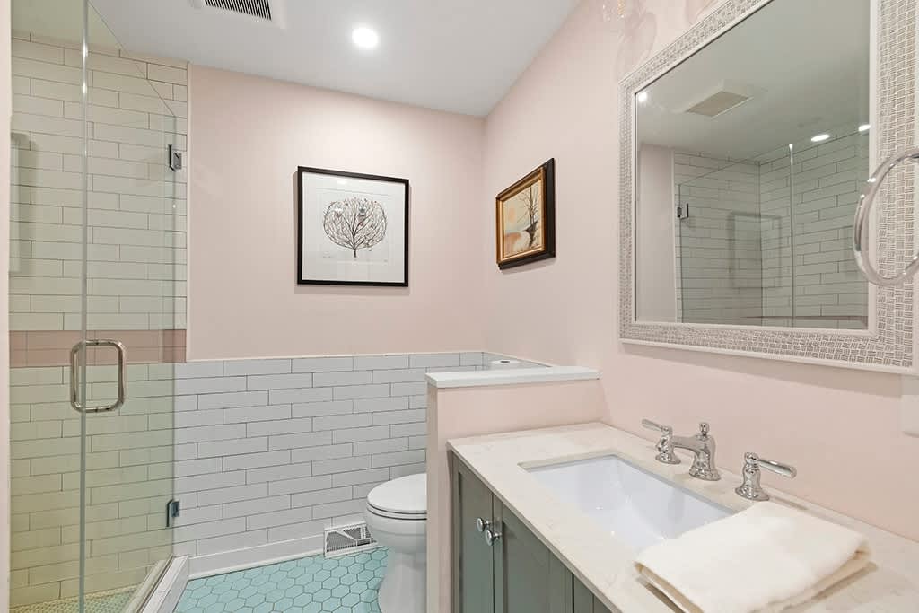 Pastel-toned bathroom with light pink walls, white subway tile wainscoting, and a glass-enclosed shower. A vanity with a white sink and a large framed mirror complete the space.
