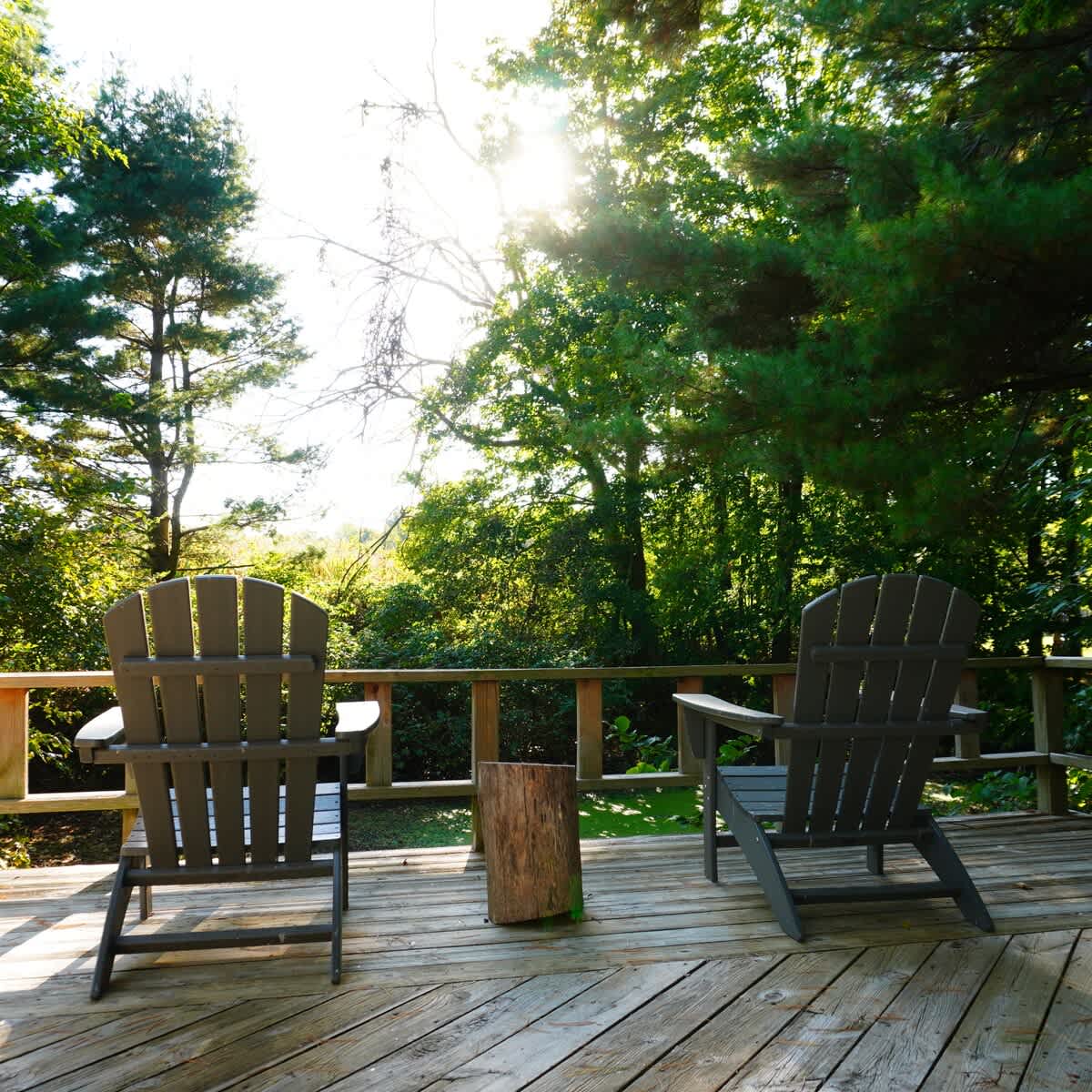 Wooden deck with two grey Adirondack chairs and a small wooden stump table, set against a backdrop of dense green trees with bright sunlight filtering through. The deck railing is visible in the foreground.