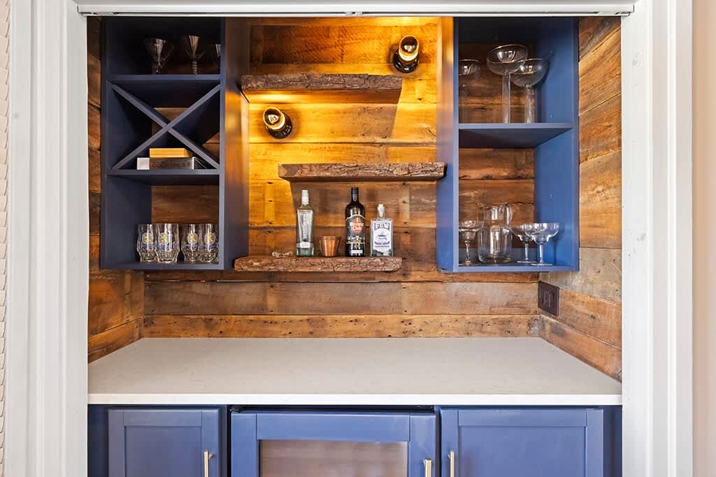 Rustic built-in bar nook with wooden plank walls, blue shelving, and live-edge wooden shelves displaying bottles and glassware. A small beverage fridge is integrated below.