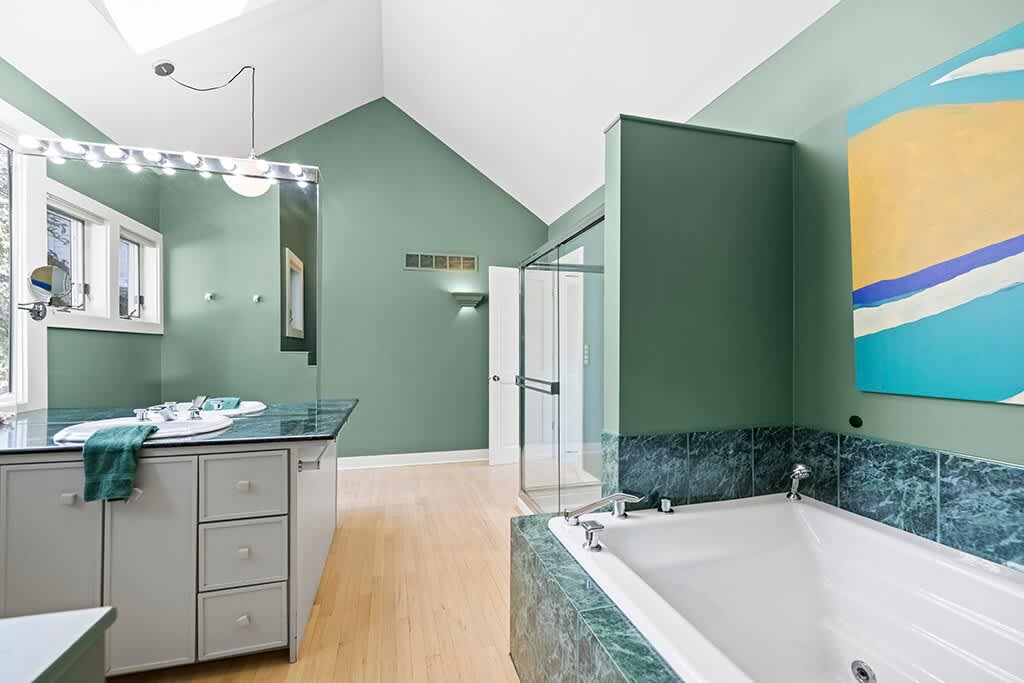 Another perspective of the spacious bathroom, highlighting the deep soaking tub with green marble surround and the adjacent glass-enclosed shower. A large abstract painting adds color to the sage green walls.