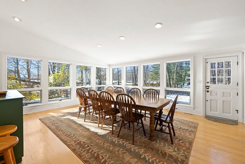 Spacious dining area featuring a large wooden dining table with ten matching wooden chairs, set on a patterned area rug. Numerous large windows offer expansive views of the snowy outdoor landscape.