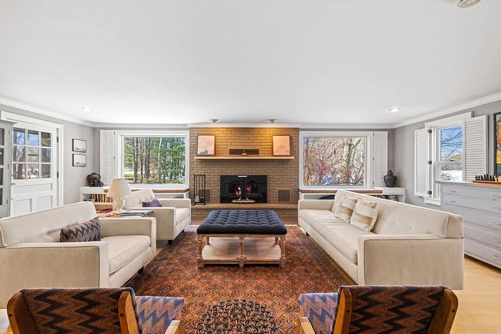 A spacious living room features light gray walls, a large brick fireplace with a wooden mantel, two light-colored sofas, two armchairs, and a dark blue tufted ottoman, all arranged on a patterned area rug.