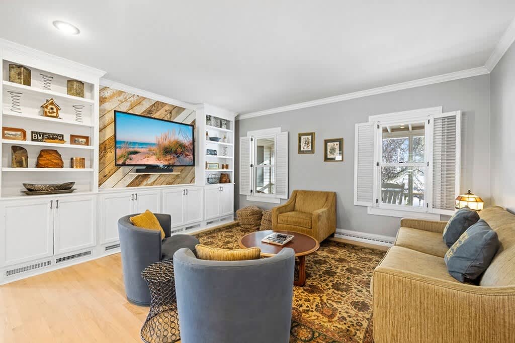 Cozy living room with light gray walls, a built-in entertainment center with a TV and shelving, and a patterned area rug. A sofa and two swivel chairs provide seating.