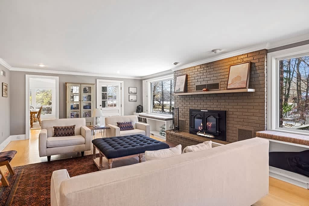 A wide-angle view of the living room highlights the brick fireplace with a burning fire, flanked by large windows with window seats, and a seating arrangement including a light sofa, armchairs, and a dark blue ottoman on a patterned rug.