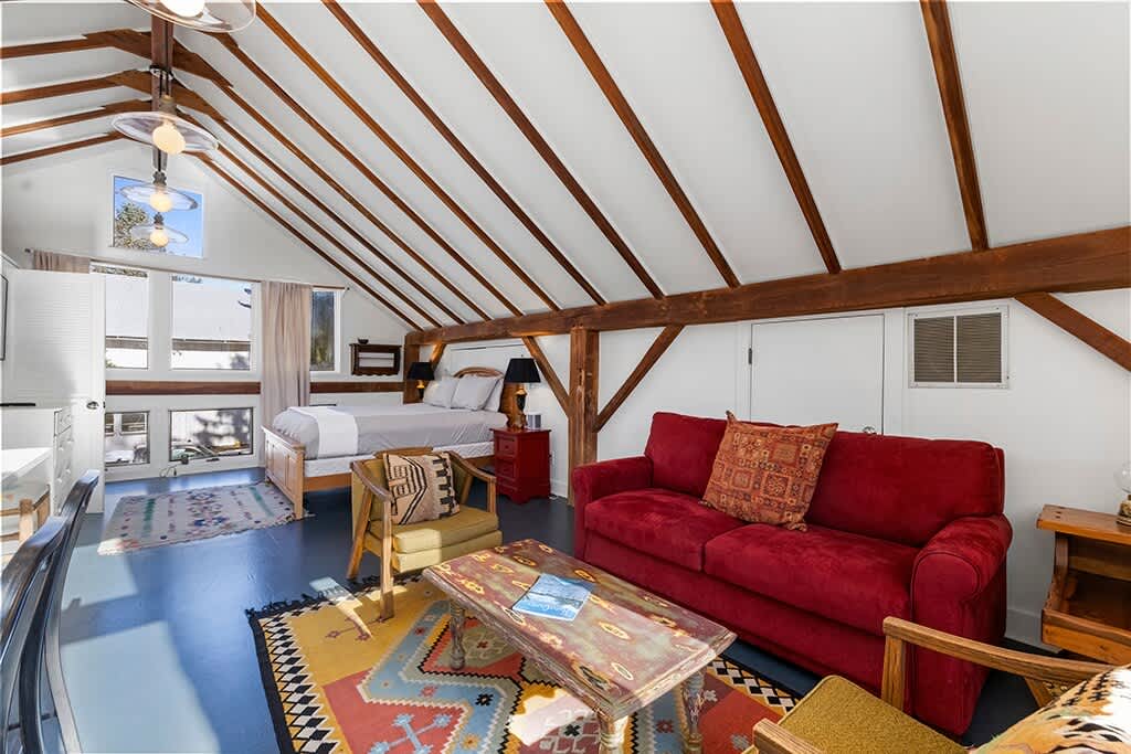 Open-plan studio space featuring a living area with a red sofa and armchairs on a colorful rug, transitioning into a bedroom area with a wooden bed frame. The room has a high vaulted ceiling with exposed wooden beams and large windows.