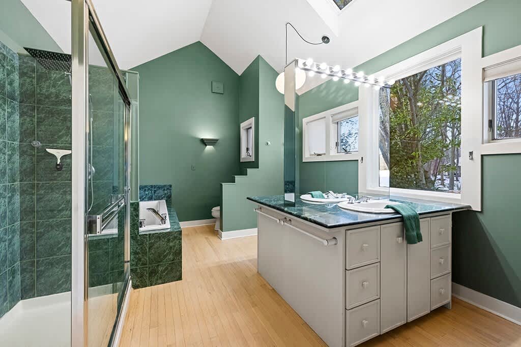 A spacious bathroom with sage green walls and a vaulted ceiling, featuring a large walk-in shower, a built-in bathtub with green marble tiling, and a double vanity with a matching green marble countertop.