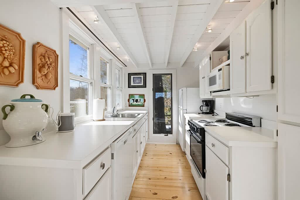 Galley kitchen with white shaker cabinets, light wood floors, and white countertops. It features a stainless steel sink, a black oven, and a white refrigerator at the far end, with an exposed beam ceiling and a view into the living/dining area.