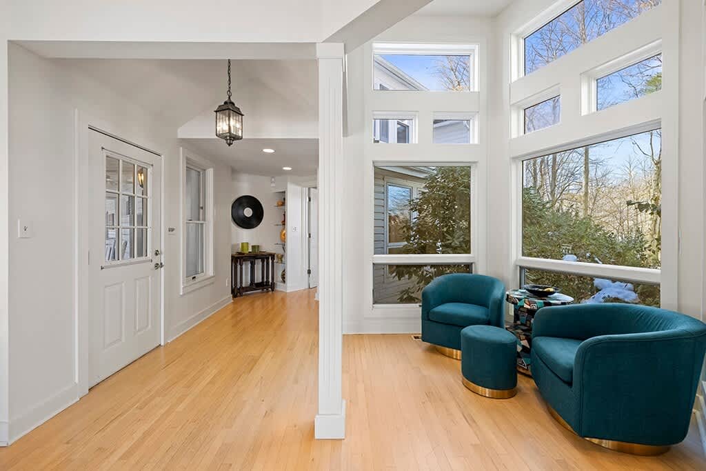 A sun-drenched sitting area features large floor-to-ceiling windows offering views of the outdoors, two teal swivel armchairs with matching ottomans, and light hardwood floors, leading into a bright hallway with a decorative console table.