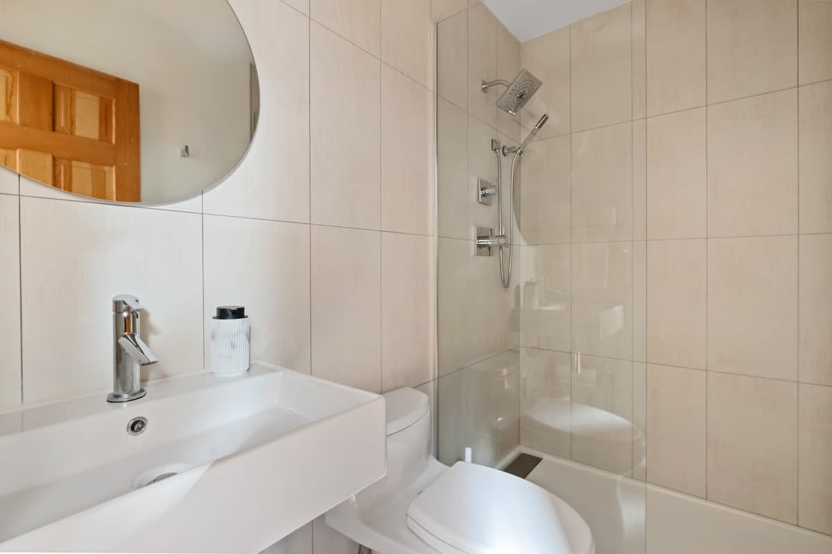 A clean, contemporary bathroom with light beige tiled walls, a white rectangular sink, a toilet, and a bathtub with a glass shower partition. A large round mirror hangs above the sink.