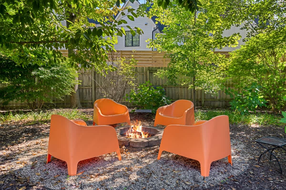 A cozy outdoor fire pit area featuring a stone fire pit surrounded by four modern orange armchairs, set on a gravel ground amidst lush green foliage and trees.