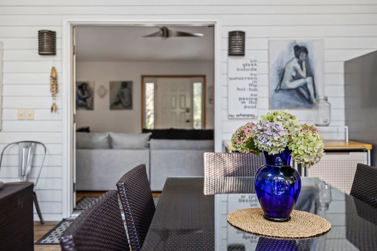 Screened-in porch with a dark wicker dining set and a blue vase with flowers on the table, offering a view into an interior living space through an open doorway.