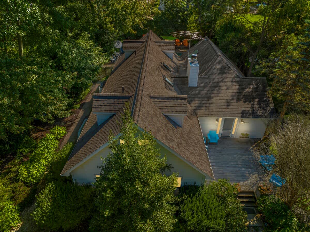 An aerial view showcasing the multi-gabled roof of a white house, surrounded by a dense canopy of green trees, with a wooden deck visible on one side.