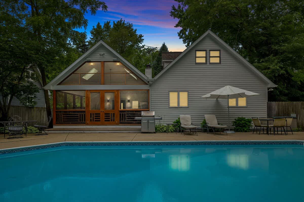 The back of a light-sided house features a screened porch with large windows and wooden doors, opening to a patio with lounge chairs and a grill, all overlooking a bright blue swimming pool at dusk.