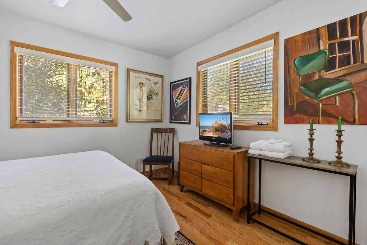 A bright bedroom with a white queen-sized bed, light hardwood floors, a wooden dresser with a television, and two windows with white blinds. Eclectic artwork and a green chair add character to the space.