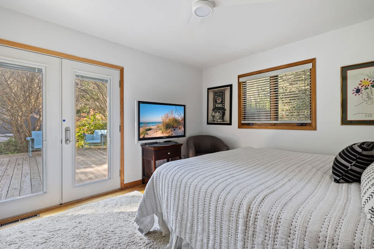 Bedroom with a queen bed, white quilted bedding, and hardwood floors. French doors open to an outdoor deck, and a flat-screen TV is placed on a wooden stand.