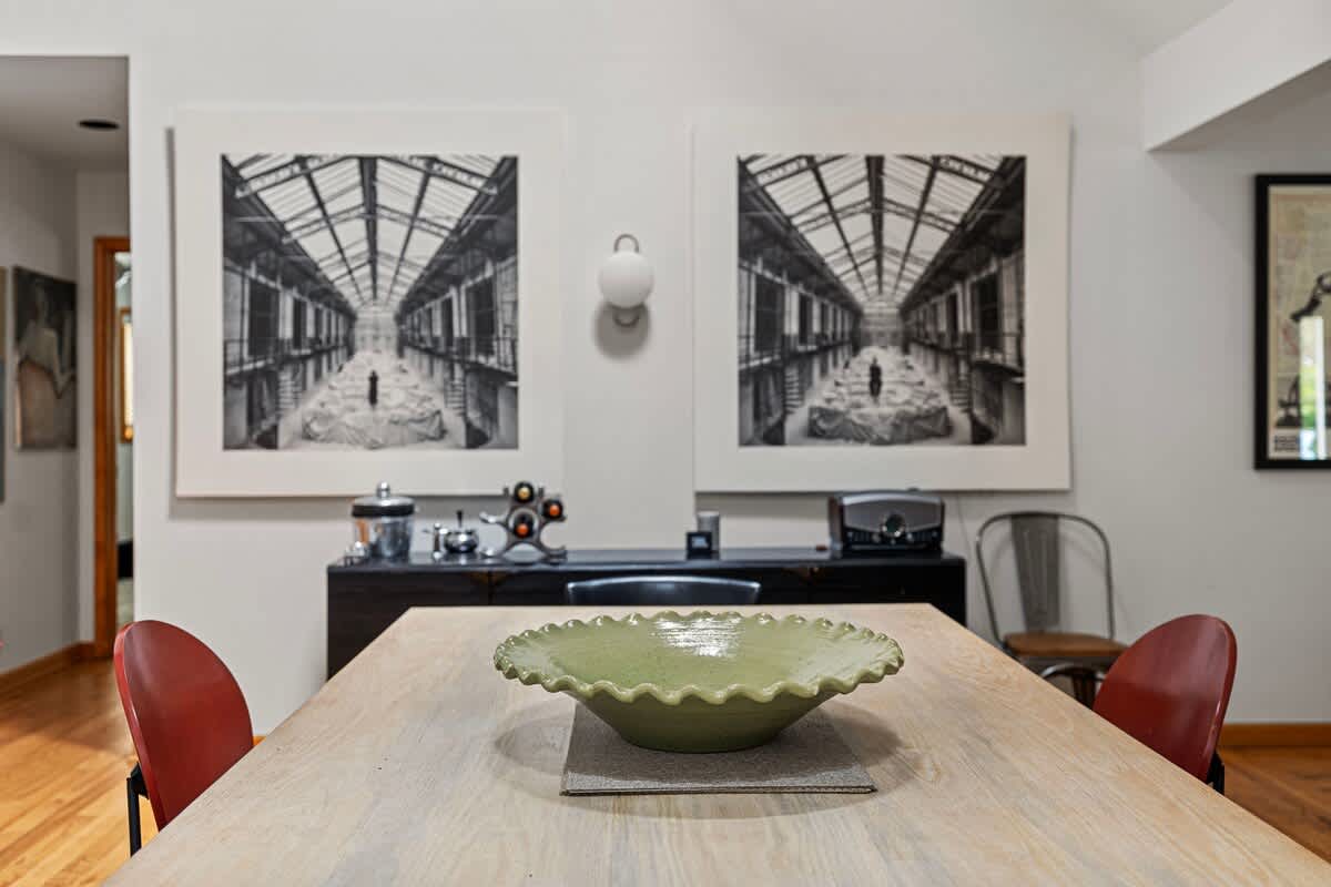 A close-up of the dining area highlights a light wooden dining table with a decorative green bowl, set against a wall adorned with two large, striking black and white architectural artworks. A dark sideboard is visible beneath the art.