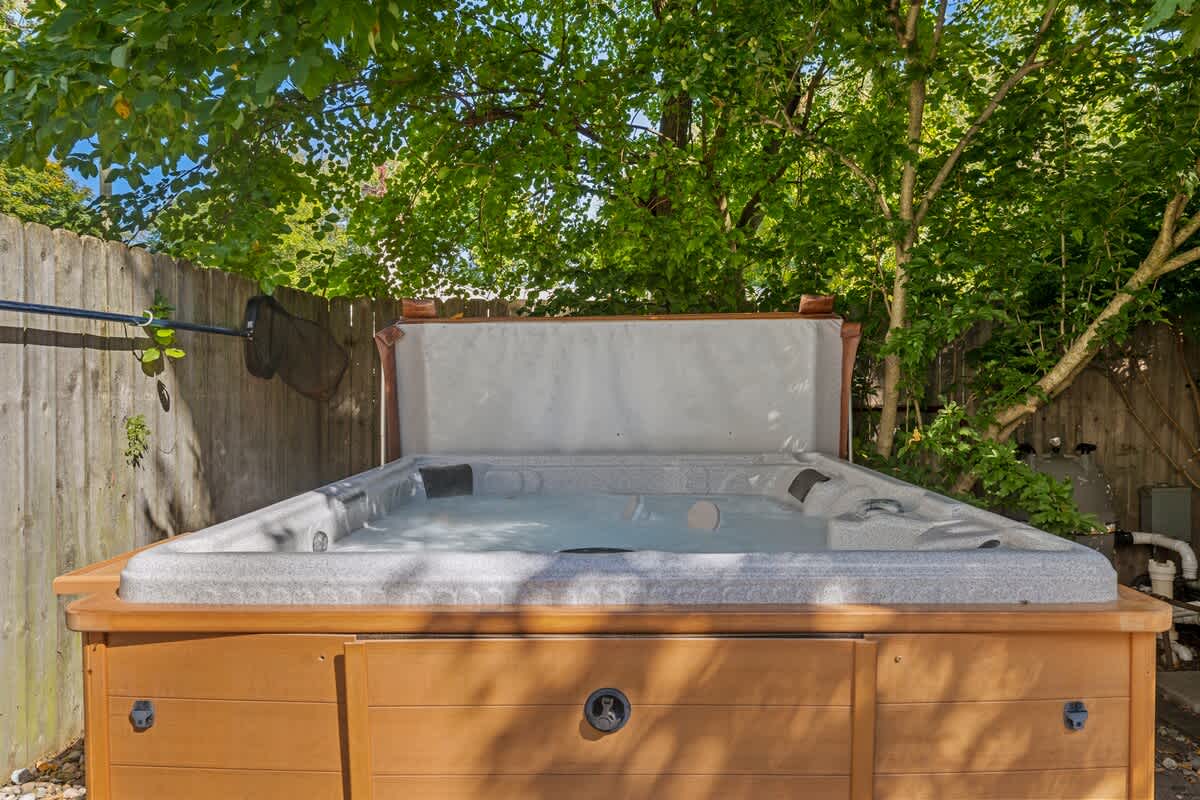A bubbling hot tub with a wooden surround and a partially open light-colored cover, nestled outdoors against a wooden fence and surrounded by green trees and foliage.