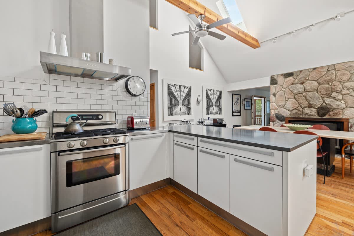Modern kitchen with white cabinets, stainless steel appliances, and a large island, opening to a dining area with a stone fireplace, all under a vaulted ceiling with exposed beams.