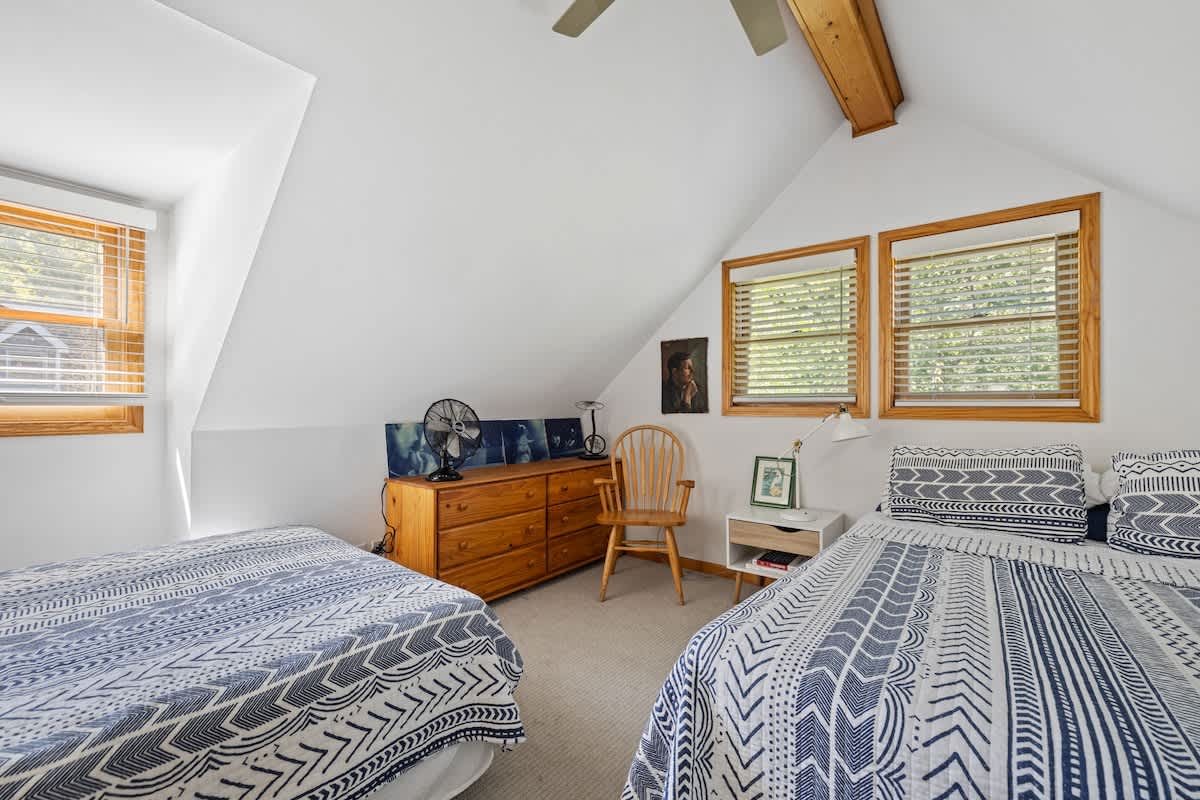 An attic bedroom featuring two twin beds with blue and white patterned bedding, a wooden dresser, and multiple windows with wooden frames and blinds. The room has a sloped ceiling and a ceiling fan.