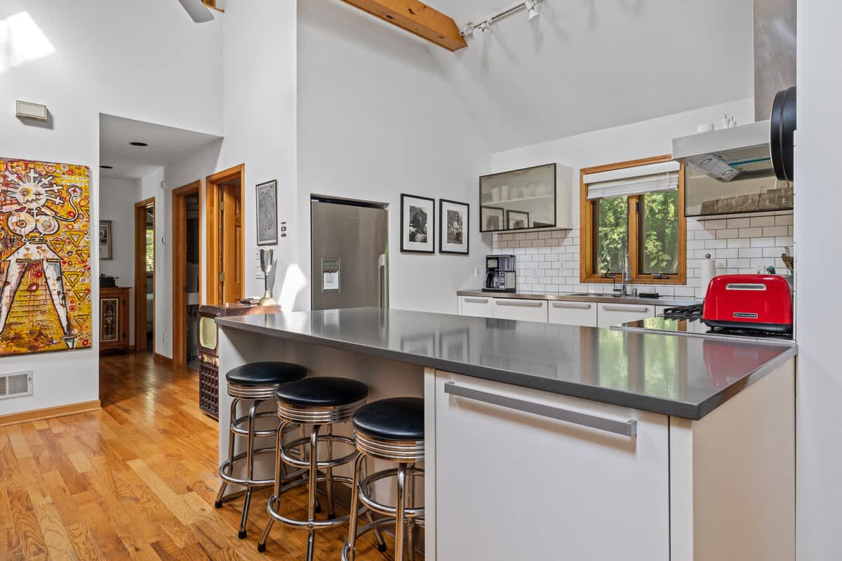 Modern kitchen with stainless steel appliances, white flat-panel cabinetry, a large island with bar stools, and hardwood flooring, leading to a hallway with wooden doors.