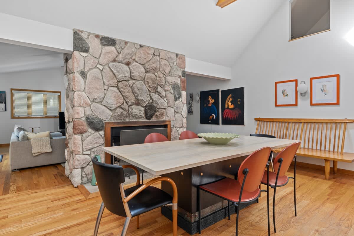 The dining area features a long light wood table with a mix of modern chairs, positioned near a large stone fireplace. Hardwood floors and vaulted ceilings contribute to the open and airy feel, with artwork visible in the background.