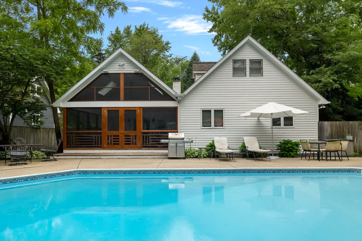 The back of the white-sided house features a prominent screened porch with large windows and a gabled roof, overlooking a sparkling blue swimming pool. A patio area with lounge chairs and an umbrella surrounds the pool, all set amidst lush trees.
