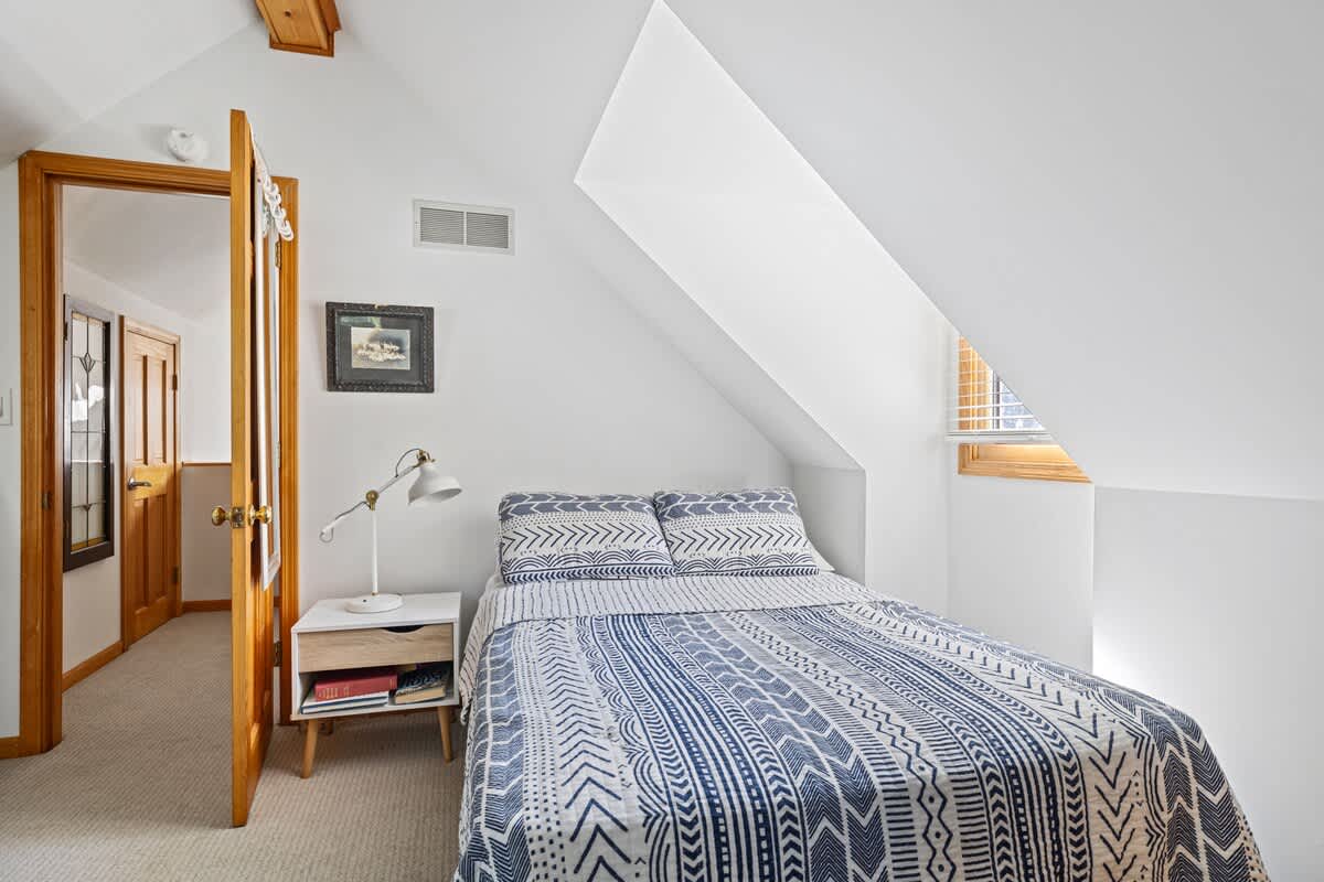 A cozy bedroom with a single bed adorned with blue and white patterned bedding, a small white nightstand with a lamp, and a sloped ceiling. A wooden door leads to a carpeted hallway.