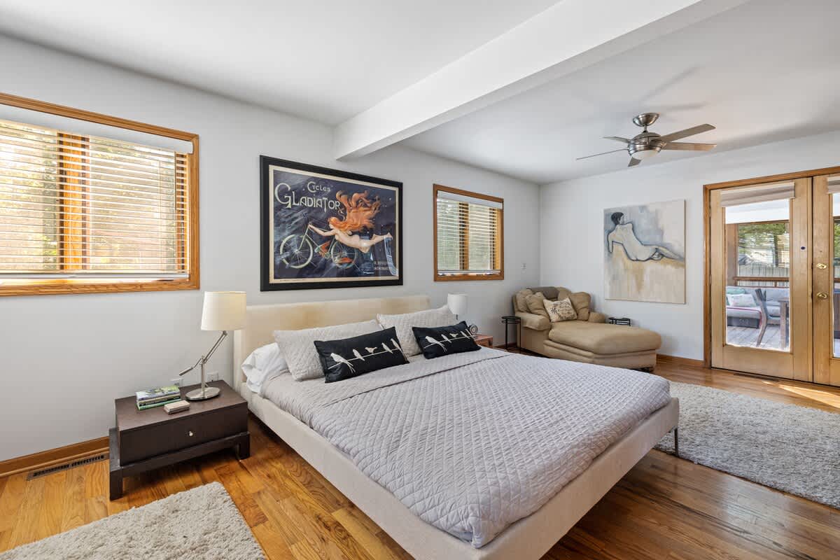 Spacious bedroom featuring a white upholstered bed with light gray bedding, hardwood floors, and white walls. A large framed art print hangs above the bed, and a chaise lounge sits in the corner.