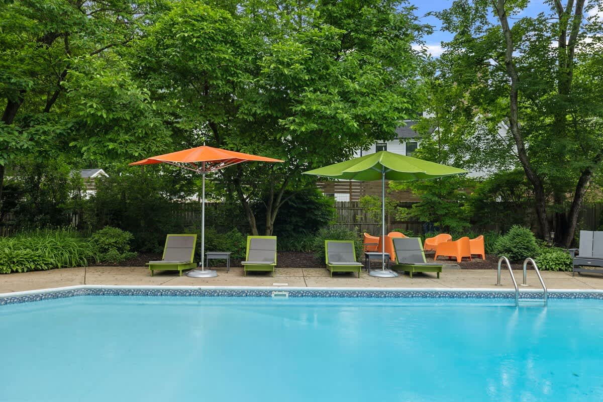 A bright blue swimming pool with a concrete deck, surrounded by green lounge chairs and colorful orange and green umbrellas, set against a backdrop of lush green trees.