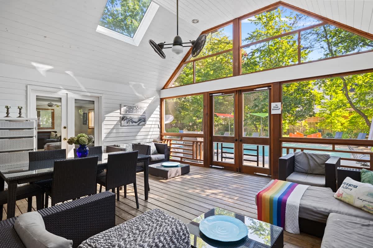 A bright, spacious screened porch features a dining table with black chairs and a lounge area with dark wicker furniture and colorful accents. Large windows and a skylight offer views of lush greenery and the outdoor pool.