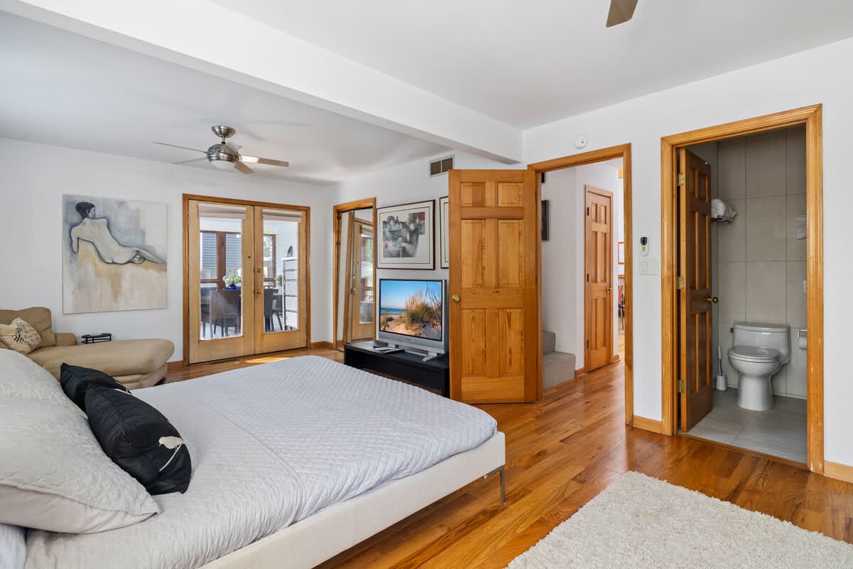 View of a bright bedroom with hardwood floors and white walls, showing a bed with light gray bedding and a TV on a low stand. French doors lead to an outdoor area, and an open doorway reveals a bathroom with a toilet.