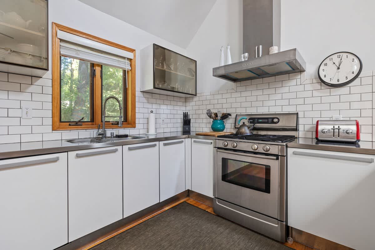 Bright kitchen with white flat-panel cabinets, stainless steel countertops, a sink under a large window, and a gas range, featuring a subway tile backsplash and hardwood floors.