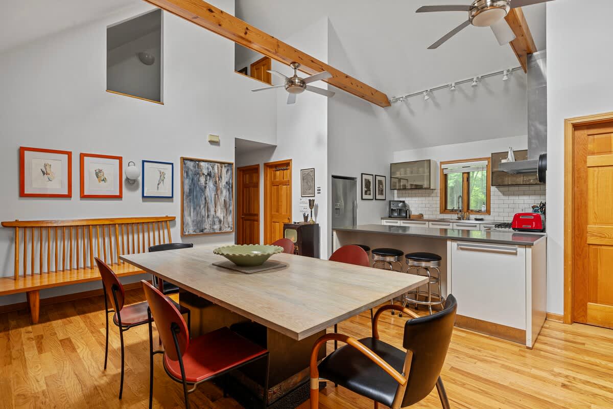 Open-concept dining area with a light wood table and mixed chairs, featuring vaulted ceilings with exposed wooden beams, and an adjacent modern kitchen with stainless steel appliances and white cabinetry.