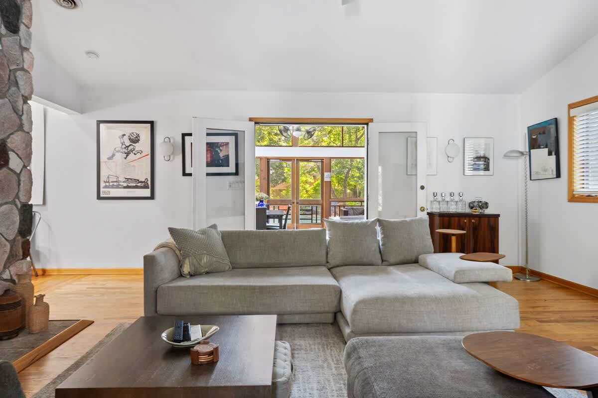 A spacious living room features a large light grey sectional sofa with throw pillows, positioned on a patterned rug. Sliding doors lead to a bright outdoor screened porch, while artwork and a wooden console table adorn the walls.