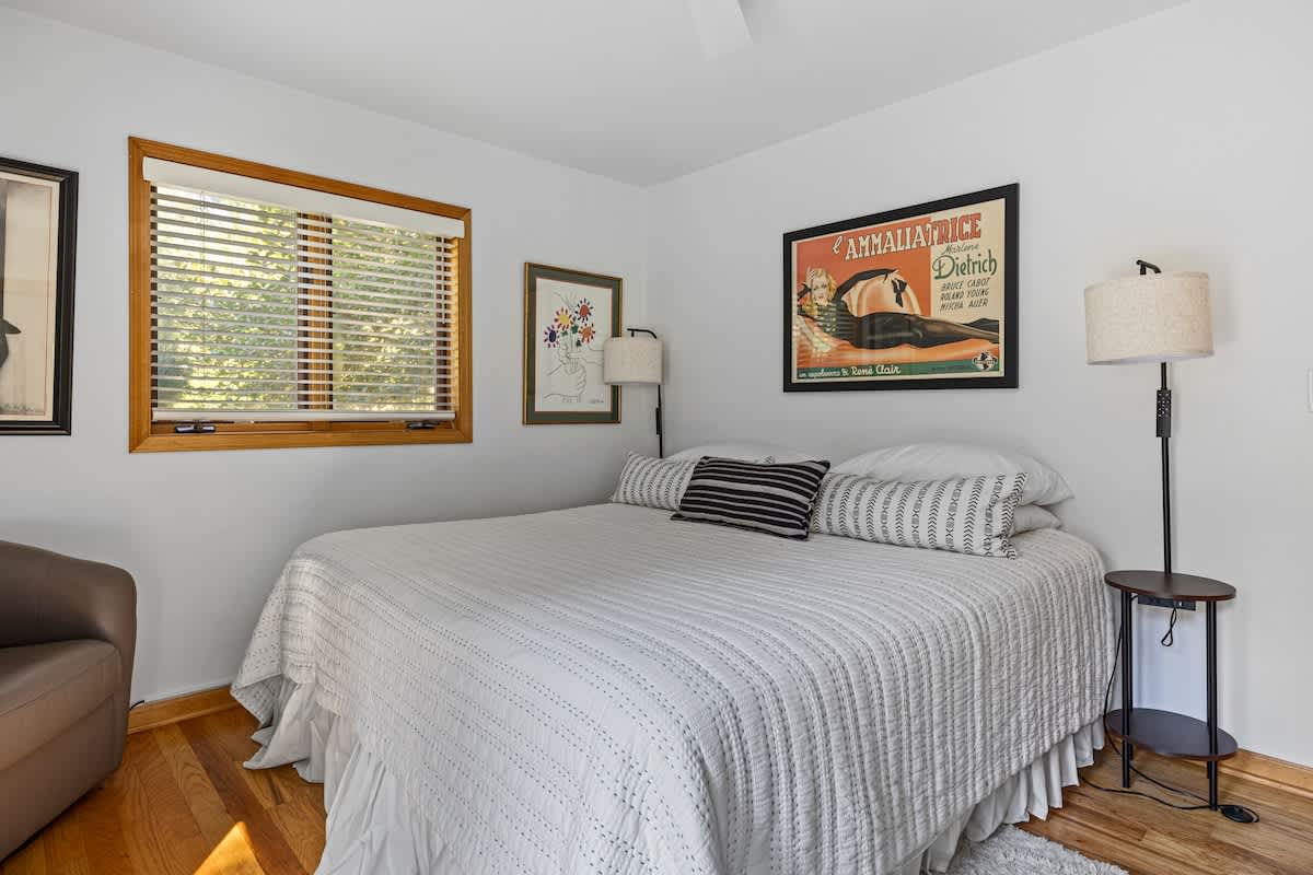 Bright bedroom with a queen-sized bed featuring white quilted bedding and black and white patterned throw pillows. Hardwood floors, white walls, and a large framed art print are visible, along with a floor lamp and a window.