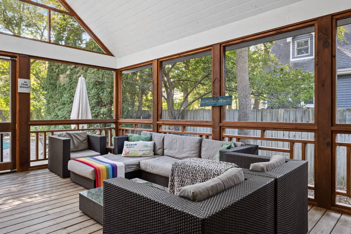 Screened-in porch with a large L-shaped outdoor sectional sofa and armchairs, a coffee table, and wooden deck flooring, offering views of the surrounding trees.