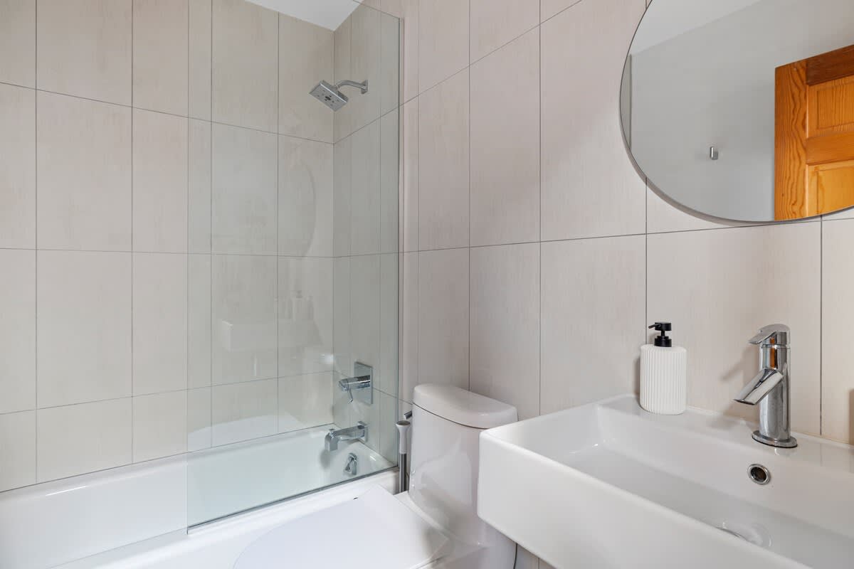 A modern bathroom featuring a white bathtub with a clear glass shower screen, a wall-mounted rectangular sink with a chrome faucet, and a toilet. The walls are clad in light beige rectangular tiles.