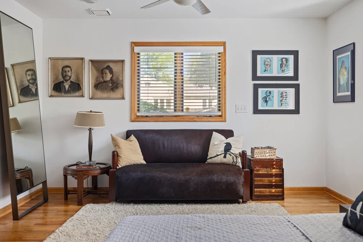Cozy corner of a bedroom with a dark brown sofa, a round wooden side table with a lamp, and framed portraits on the white wall. A large full-length mirror leans against the wall, and a window with wooden blinds provides natural light.