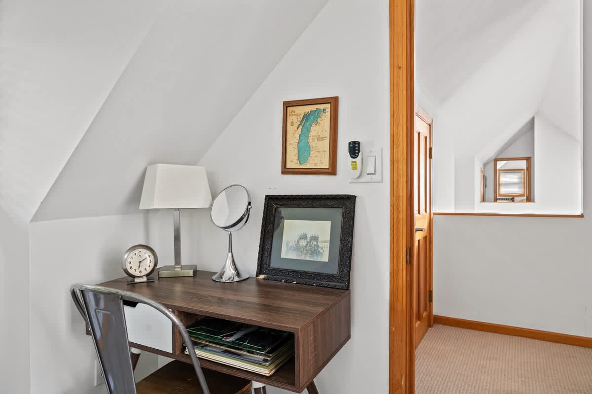 A dedicated workspace nook with a dark wood desk, a white table lamp, and a round vanity mirror, set against a white wall with a framed map. The area has a carpeted floor and a sloped ceiling.