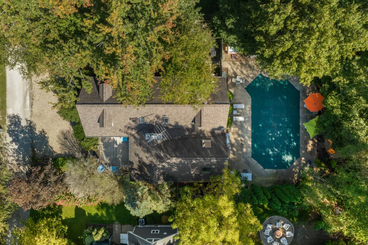 A high aerial view of the property, revealing a house with a brown roof, a covered swimming pool, and a fire pit area, all nestled within a vast expanse of lush green trees.