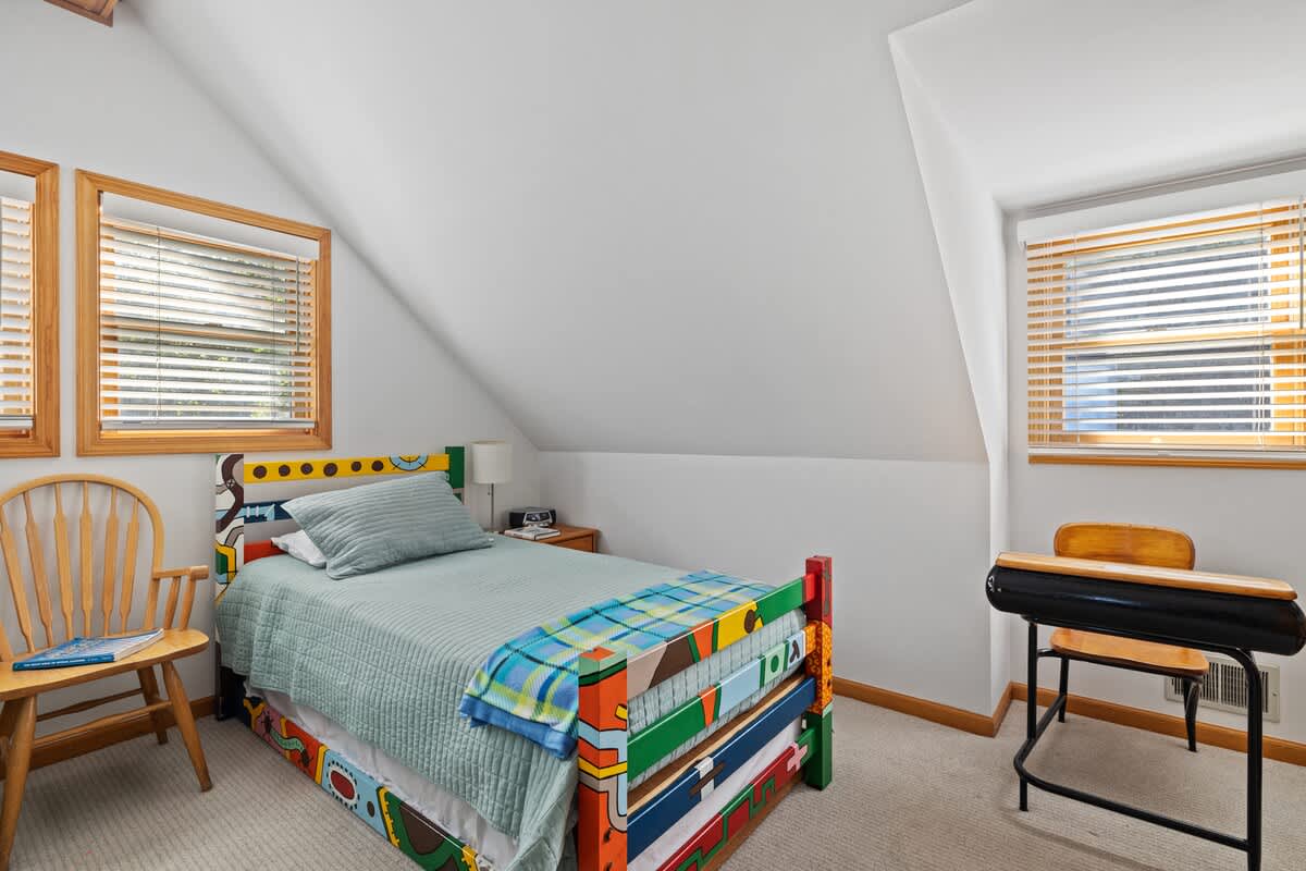 A cheerful corner of a bedroom featuring a single bed with a colorful painted headboard and light green bedding, alongside a small vintage-style school desk. A window with blinds provides natural light.