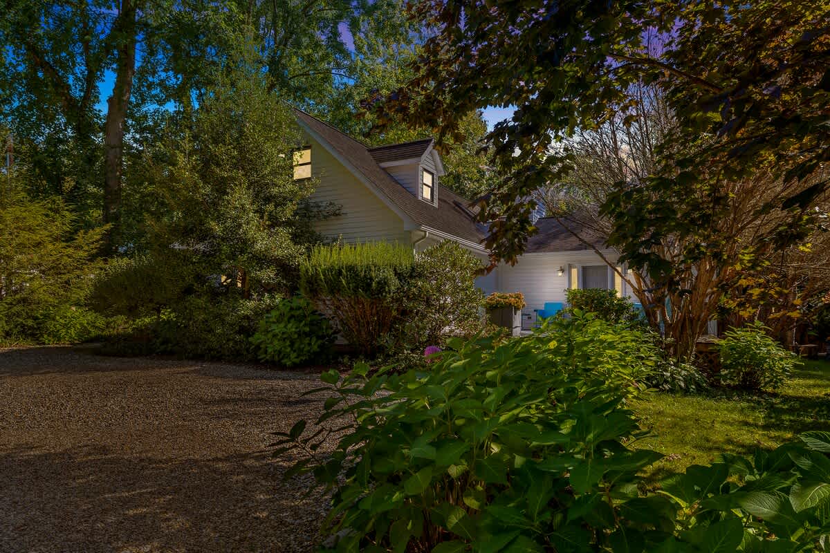 A charming white house with a gabled roof, illuminated at dusk with interior lights glowing, surrounded by lush green trees and shrubs, with a gravel driveway in the foreground.