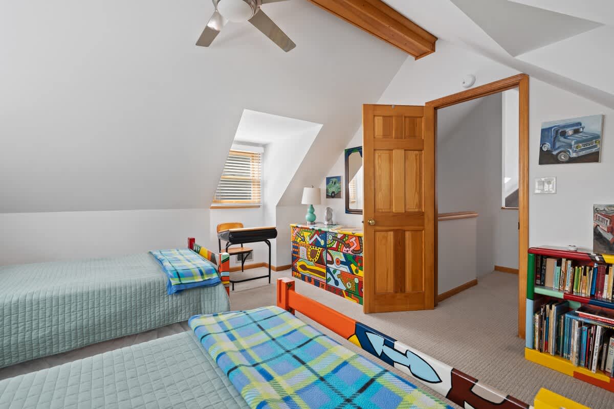 A spacious attic bedroom with two single beds featuring colorful painted headboards, a matching colorful dresser, and a vibrant bookshelf filled with books. The room has a sloped ceiling and a ceiling fan.